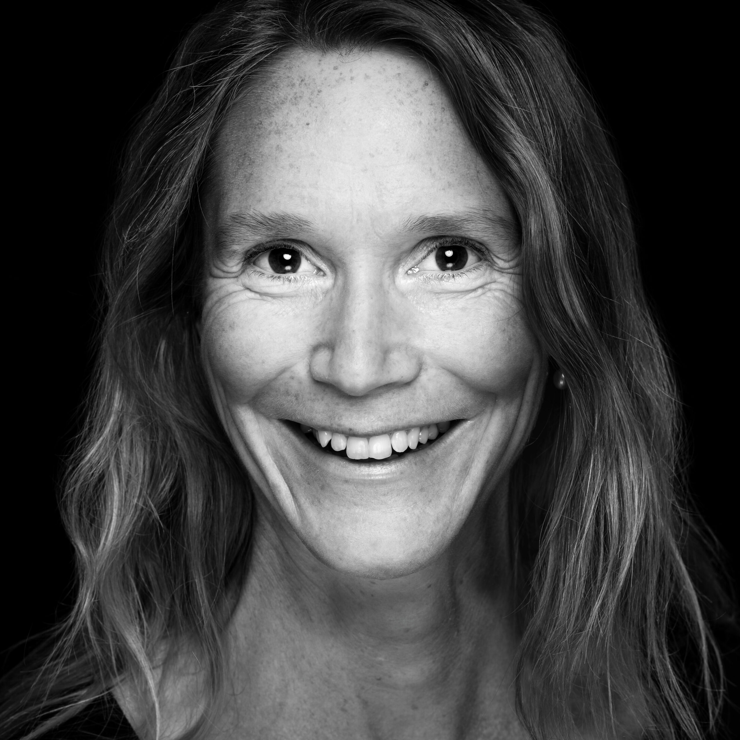 Das Bild zeigt die Autorin Corinne Sutter in einem ausdrucksstarken Schwarz-Weiß-Porträt. Sie lächelt herzlich und blickt direkt in die Kamera, was ihr Gesicht offen und freundlich erscheinen lässt. Ihr langes, welliges Haar umrahmt ihr Gesicht natürlich. Das dunkle Hintergrund kontrastiert stark mit ihrem Gesicht und betont ihre lebendigen Augen und das fröhliche Lächeln. Die Aufnahme strahlt Wärme und Authentizität aus und lenkt den Fokus ganz auf ihre Ausstrahlung.