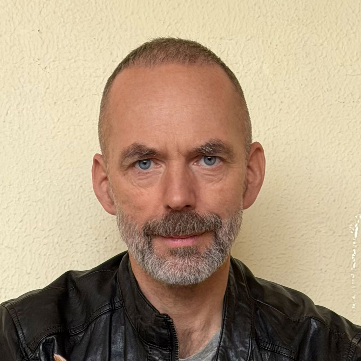 Porträt des Autors Marc Ebersbach. Er blickt in die Kamera, hat kurzes Haar und einen grauen Bart und trägt eine schwarze Lederjacke vor einer hellen, neutralen Wand.