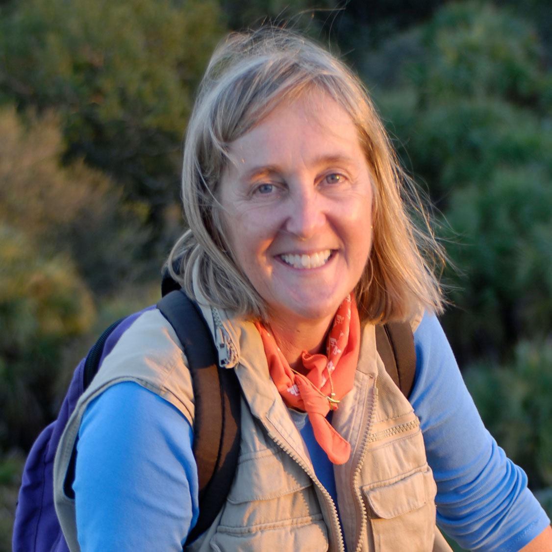Porträt der Autorin Meg Lowman im Freien. Sie lächelt in die Kamera, trägt ein hellblaues Oberteil, eine beige Weste, ein orangefarbenes Halstuch und einen Rucksack; im Hintergrund ist unscharfe Natur zu sehen.