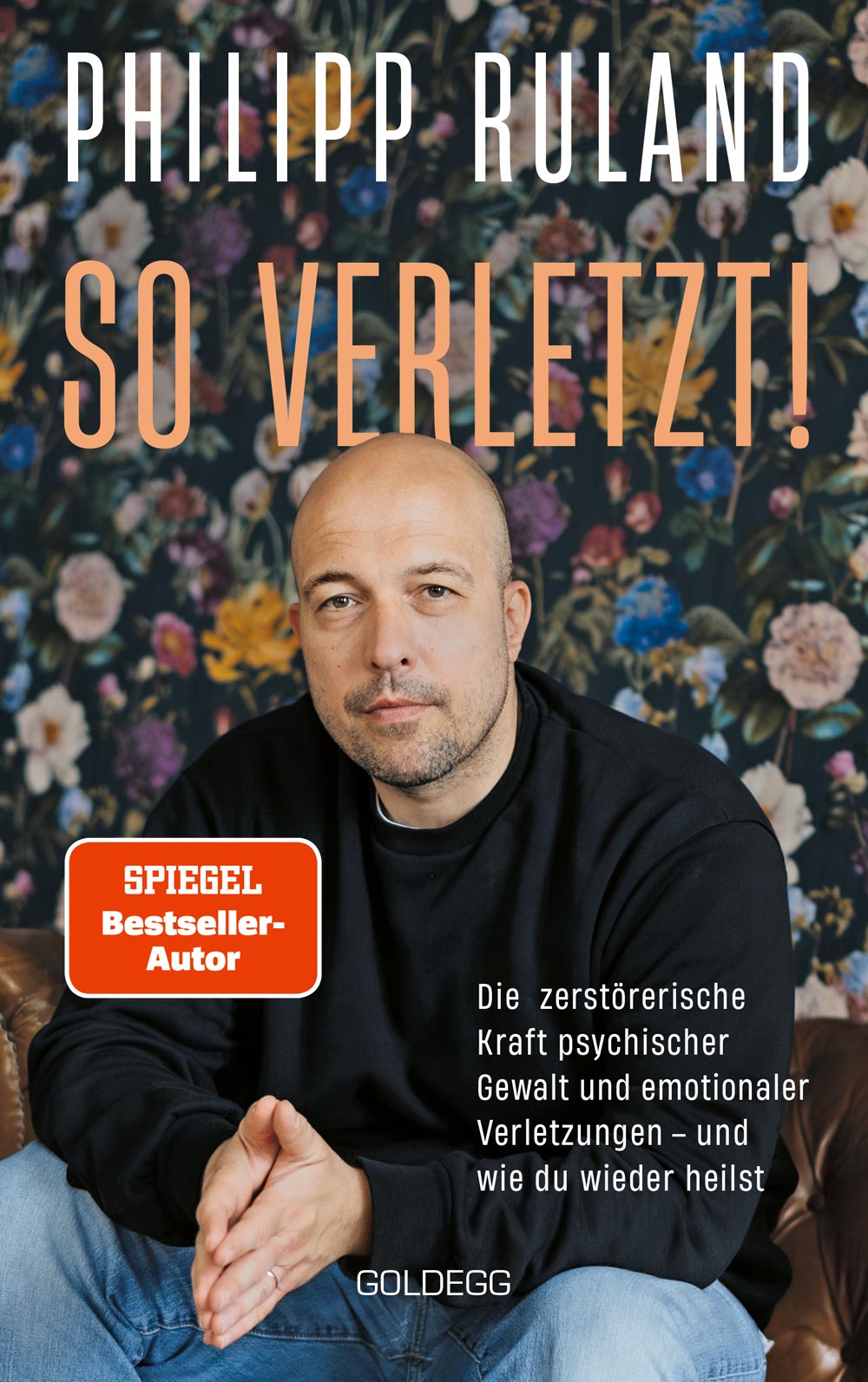 Buchcover von So verletzt! von Philipp Ruland. Auf dunklem, floral gemustertem Hintergrund sitzt eine nachdenklich blickende Person in schwarzem Pullover auf einem Ledersessel. Der Titel steht groß in apricotfarbener Schrift im oberen Bereich. Links ist ein rotes Label mit dem Hinweis ‚SPIEGEL Bestseller-Autor‘. Rechts unten befindet sich der Untertitel ‚Die zerstörerische Kraft psychischer Gewalt und emotionaler Verletzungen – und wie du wieder heilst‘. Unten ist das Goldegg-Verlagslogo abgebildet.